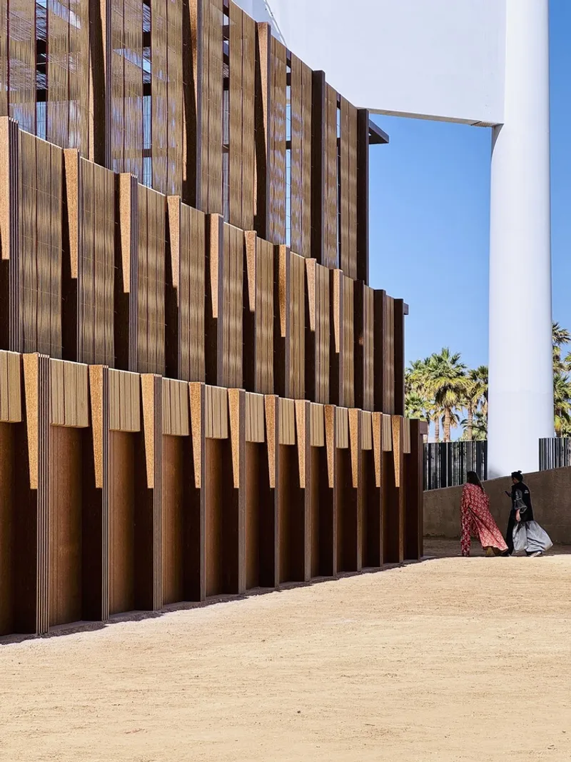 Vertical timber screen facade with staggered slats as two figures walk past in traditional dress