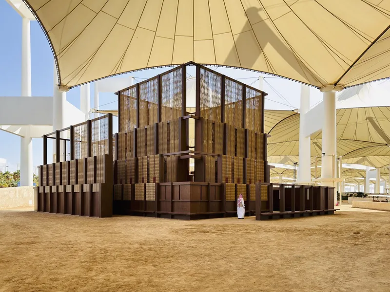 Stacked timber-clad volumes beneath a large fabric canopy with white columns and sandy ground