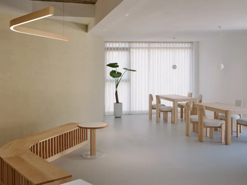 Open interior with blonde wood furniture, a potted plant and sheer curtains filtering daylight