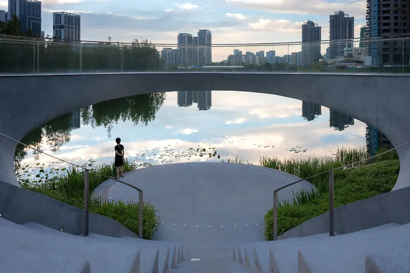 Curved concrete staircase descending to a pond with planted grasses and a figure at dusk