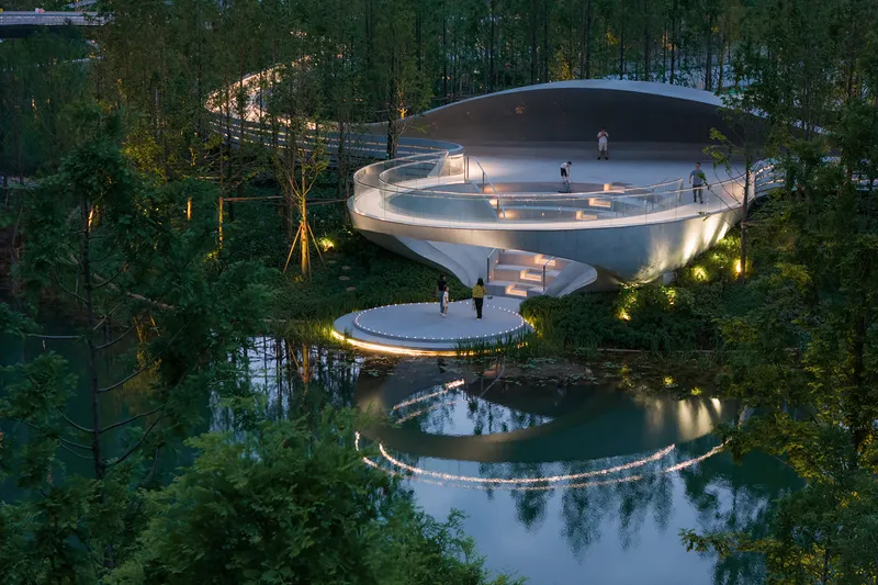 Spiraling white concrete structure with illuminated edges and visitors reflected in the pond below at twilight