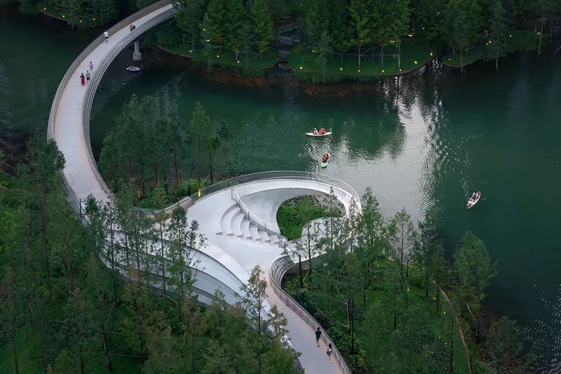 Aerial view of curving white amphitheater and pathway along a forested lake with kayakers on the water