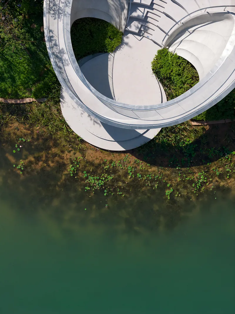 Top-down view of spiraling white walkway wrapping around circular planted voids above shallow water