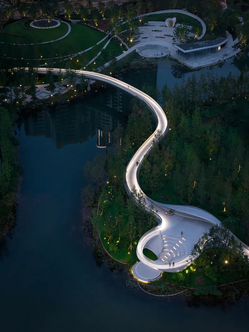 Evening aerial view of illuminated curved pathways and amphitheater threading through forested islands and water