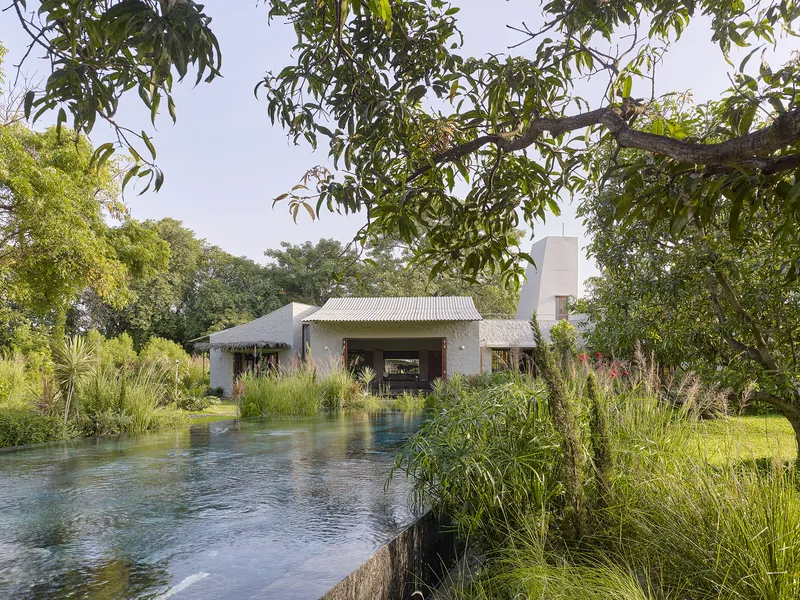 White rendered pavilion with corrugated metal roof seen across a planted water pool surrounded by grasses