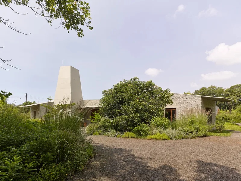 Long view of the low-slung residence with tapering chimney tower rising above planted gravel courtyard
