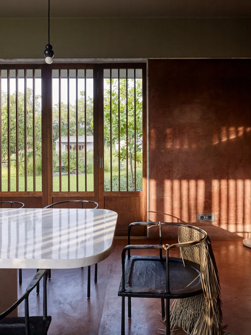 Dining area with vertical timber screen framing garden views and striped shadow patterns on terracotta plaster wall