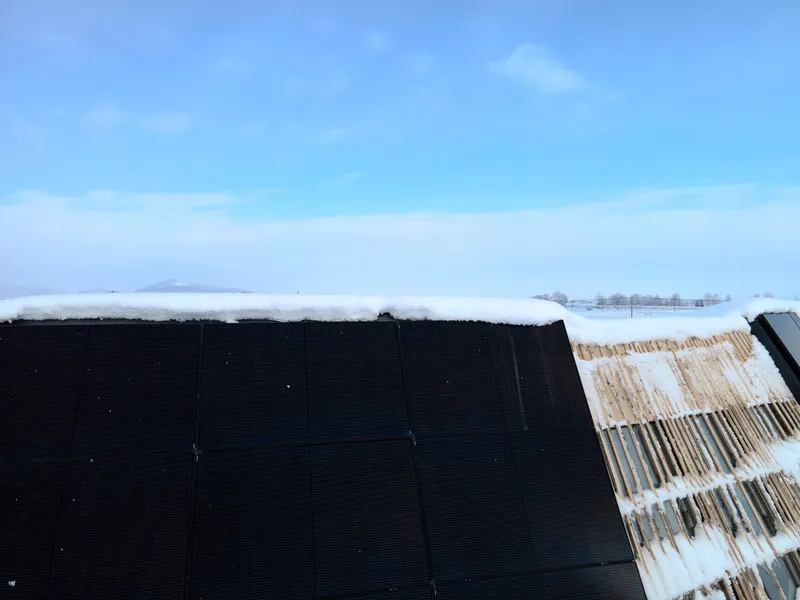 Roofline junction showing black corrugated metal meeting vertical timber slats with hills in the distance
