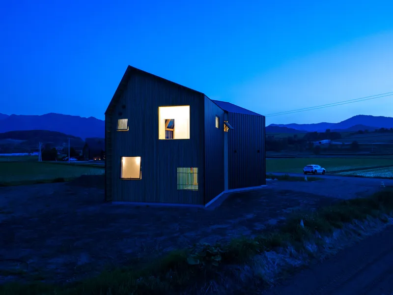 Black corrugated metal house with illuminated windows at dusk in a rural landscape
