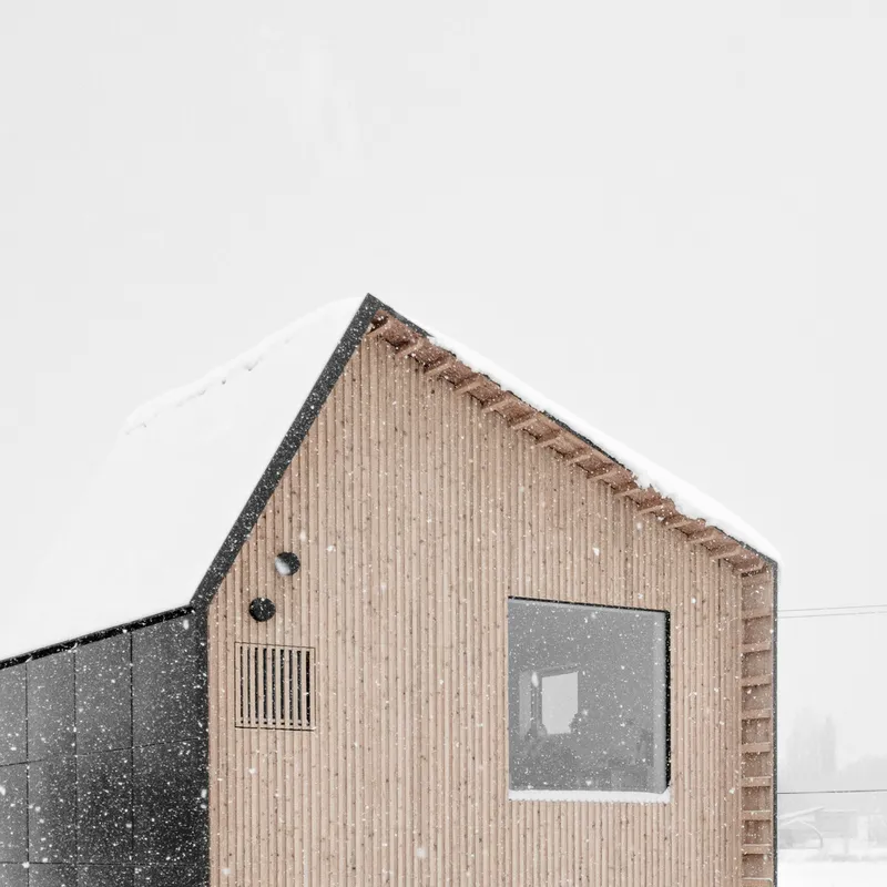 Gabled facade with vertical timber cladding, square window and louvered vent panel during snowfall
