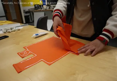The image shows a person fixing two pieces of orange fabric together using liquid stitch. The fabric