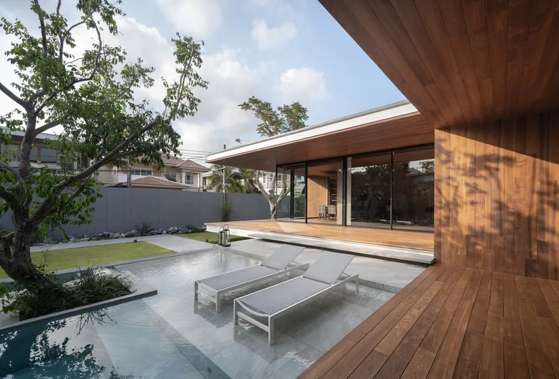 Shallow water feature with submerged loungers on deck beneath cantilevered timber soffit and dappled tree shadows