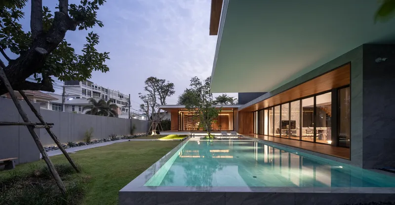 Swimming pool stretching along glazed living spaces at twilight with courtyard trees silhouetted against sky