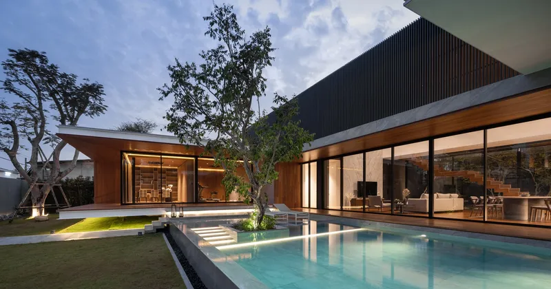 Rear courtyard with illuminated pool and glass-walled volumes framed by mature trees at dusk