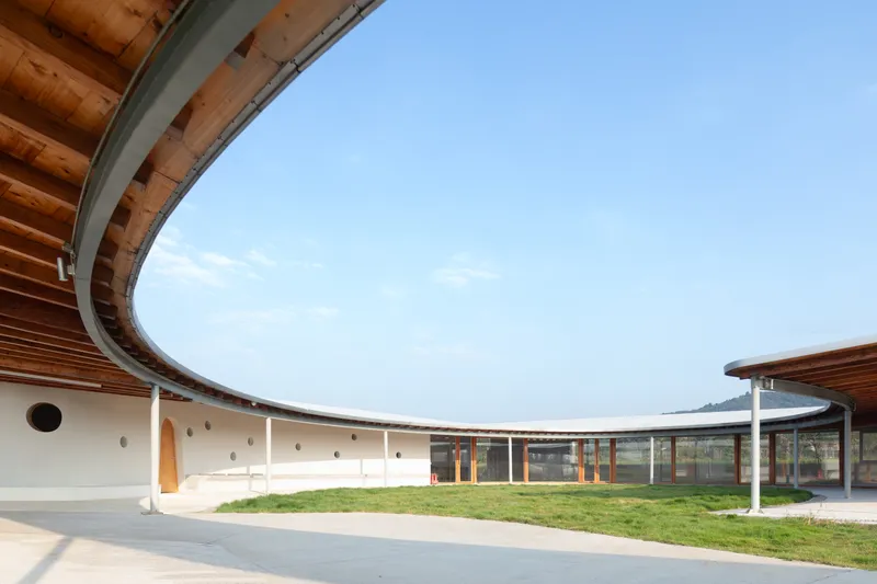 Curved facade with timber columns and overhanging roof wrapping around a central lawn under clear sky