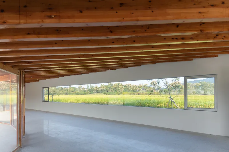 Interior gallery space with horizontal window framing rice fields beneath exposed timber beam ceiling
