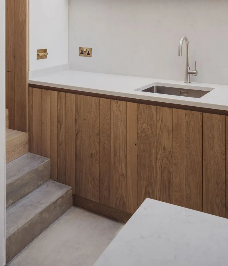Oak kitchen cabinetry with white countertop and undermount sink beside concrete stairs
