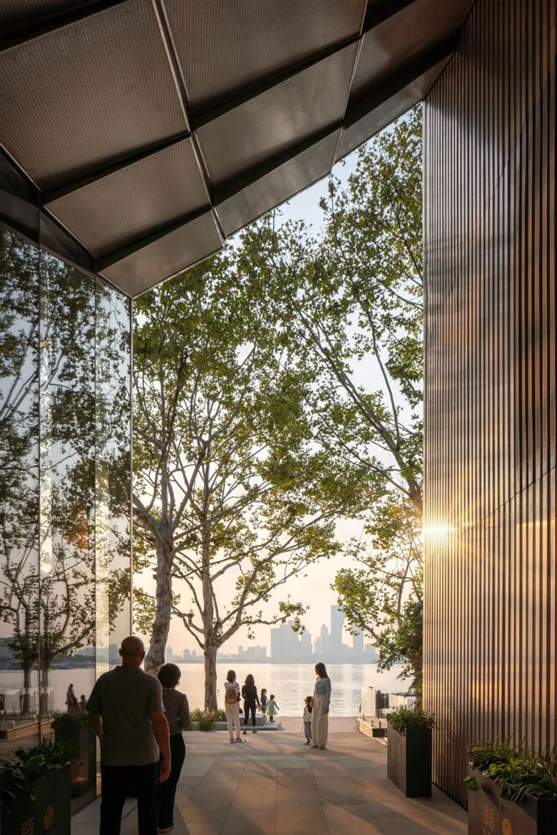 View through glass wall to waterfront terrace with visitors gathering beneath mature trees at dusk