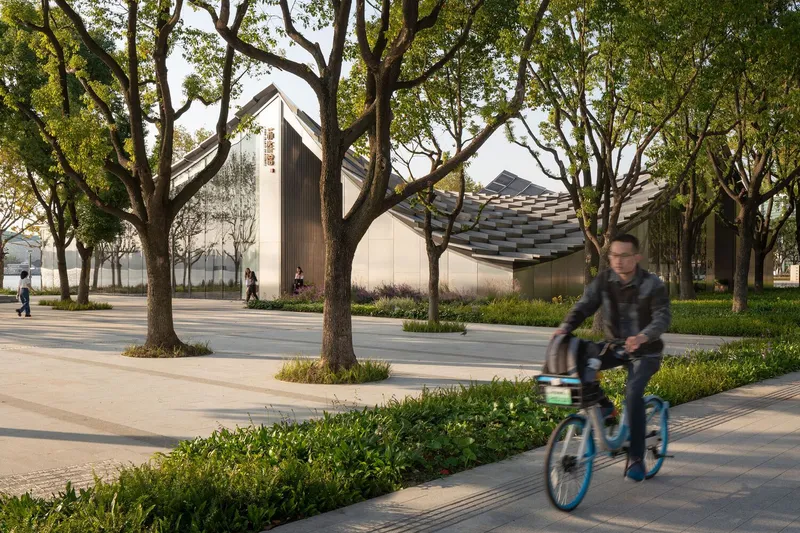 Exterior view of the curved roof volume seen through a grove of mature trees with cyclist passing