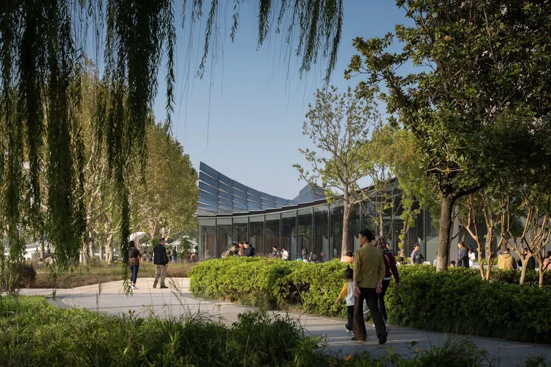 Glazed pavilion with curved solar panel roof set among willows and pathways with visitors strolling