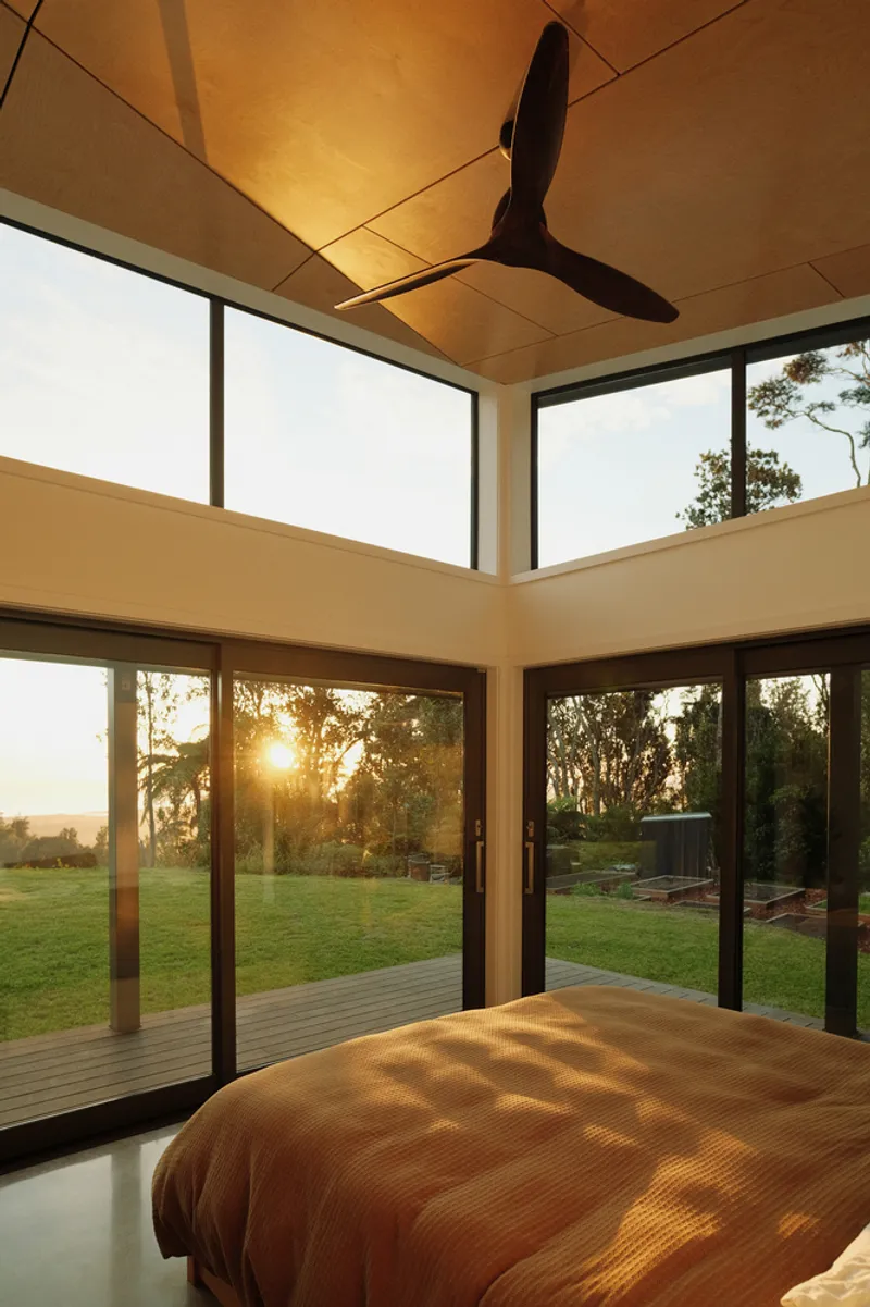 Interior bedroom corner with plywood ceiling panels and full-height glazing overlooking lawn at sunset