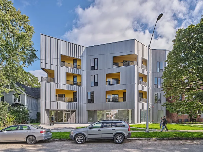 Street view of the four-story vertical metal panel facade with yellow balconies framed by green trees