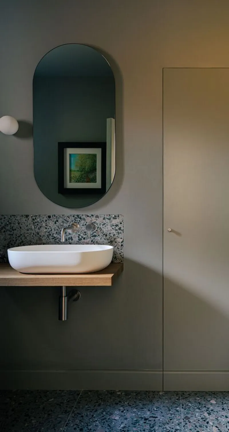 Bathroom vanity with terrazzo backsplash, floating timber shelf, and arched mirror reflecting framed artwork