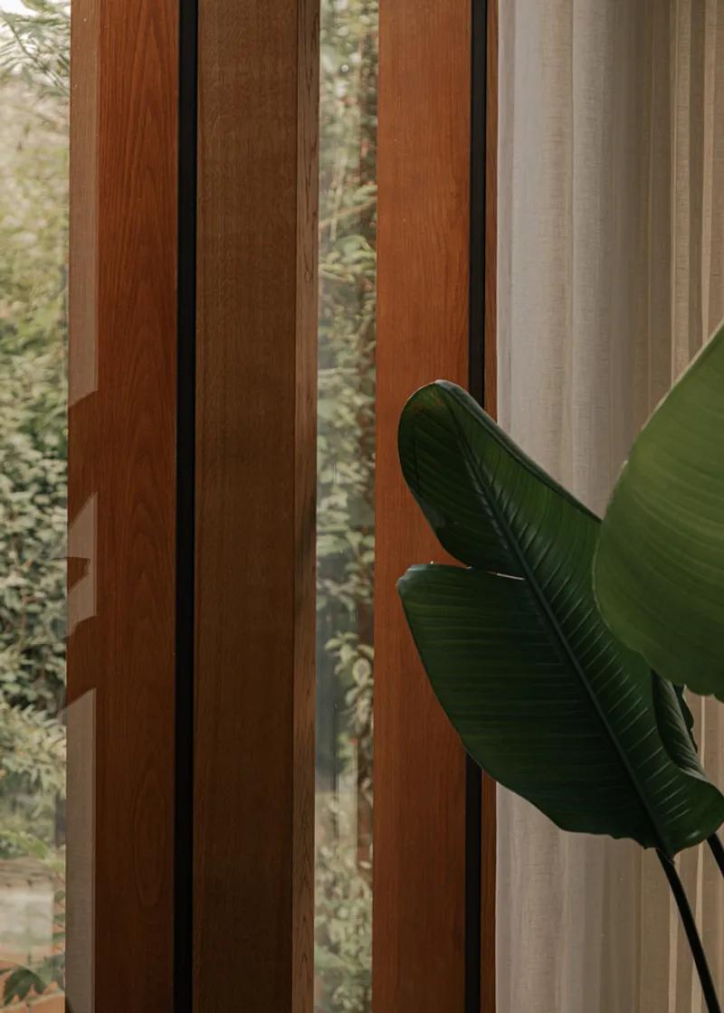 Vertical timber mullions with dark metal joints beside a banana leaf reaching into the frame