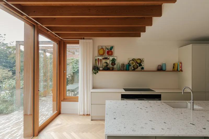 Kitchen island with terrazzo countertop beneath timber beams and corner glazing to garden