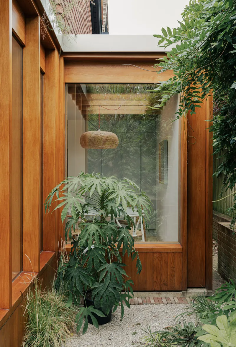 Timber-framed entrance with vertical cladding and glazed door opening to a planted courtyard with foliage
