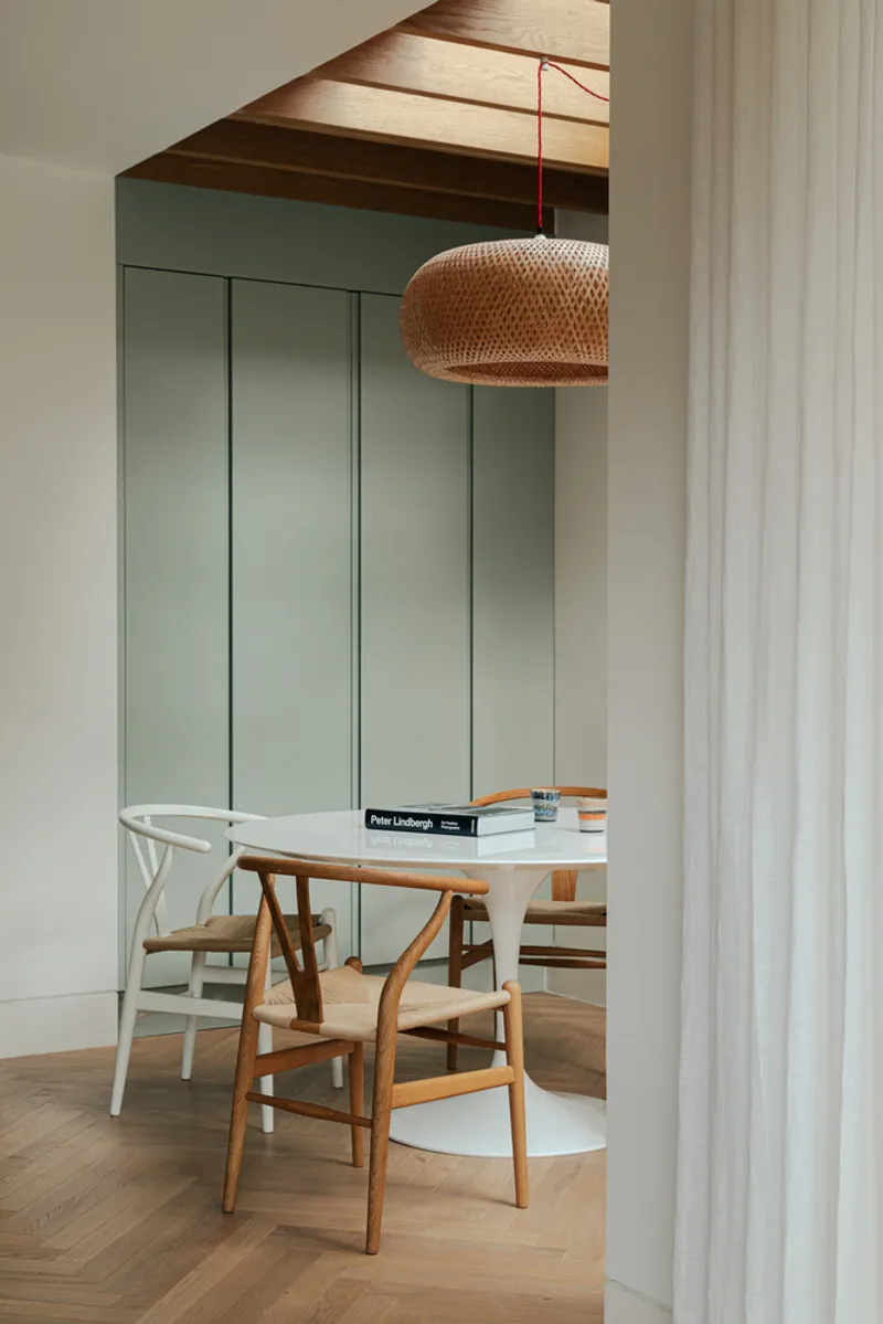 View through sheer curtain to dining table under woven pendant light with green paneled wall beyond