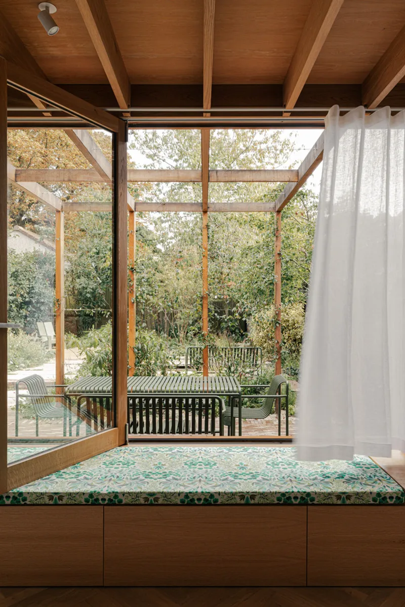 Interior window seat with cushioned bench facing glazed timber doors and outdoor dining beneath a covered pergola