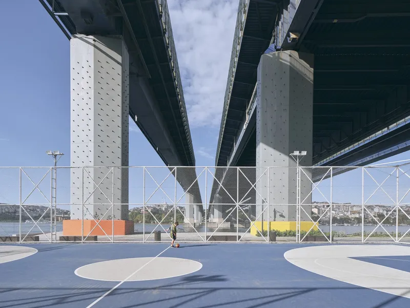 Sports court surface with white painted markings beneath concrete bridge piers and metal mesh barriers