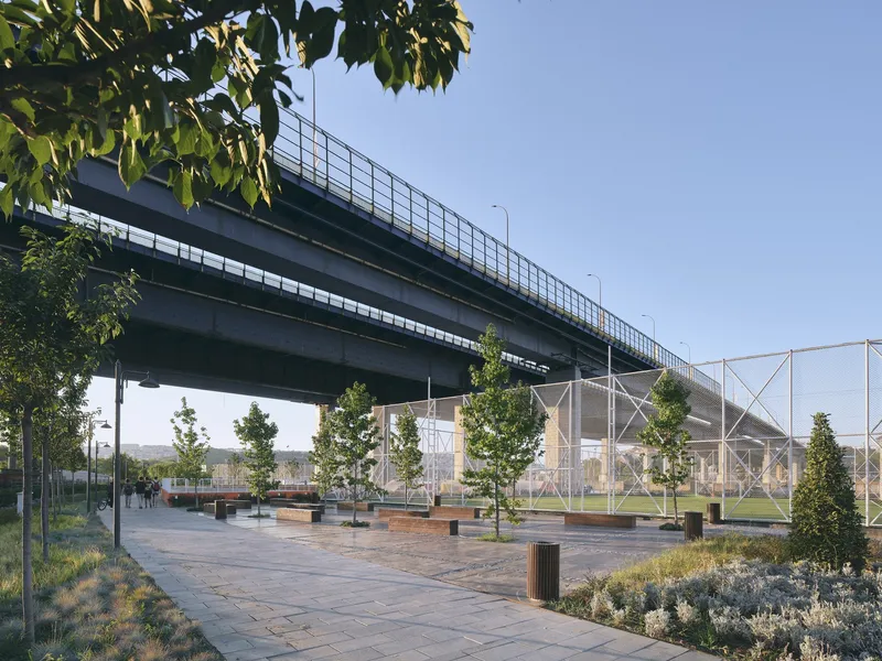 Paved plaza beneath highway bridge with tree-planted beds and metal mesh enclosure at dusk