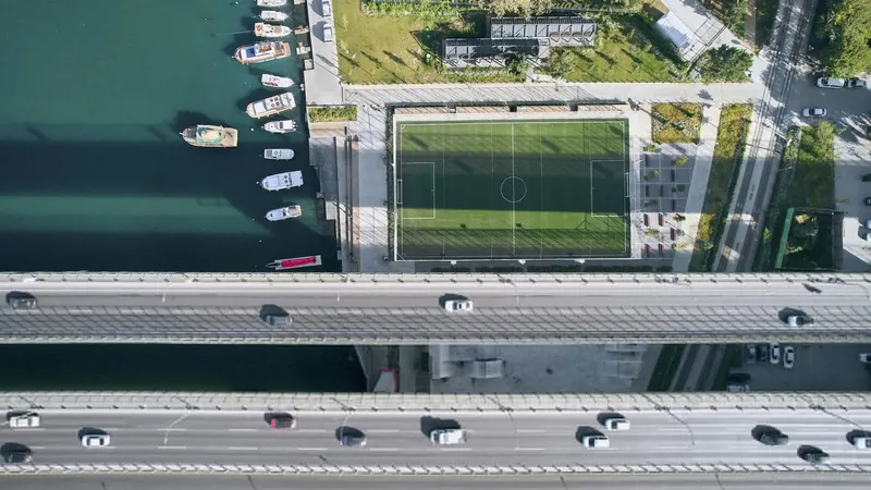 Aerial view of soccer field between highway bridge deck and waterfront with moored boats