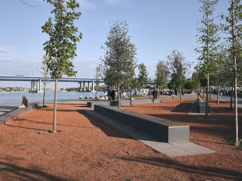 Waterfront plaza with red gravel surface, concrete benches, and young trees overlooking a river and bridge