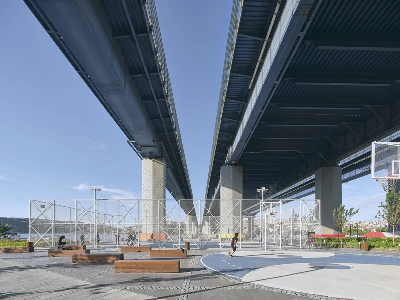 Basketball court beneath twin highway bridges with metal mesh fencing and concrete piers under clear blue sky