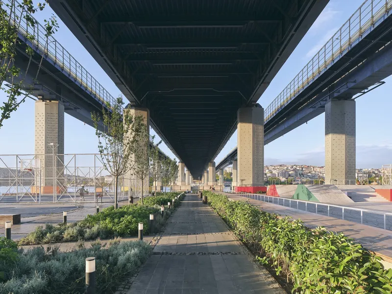 Paved pedestrian path beneath highway bridges flanked by planted beds with shrubs and young trees