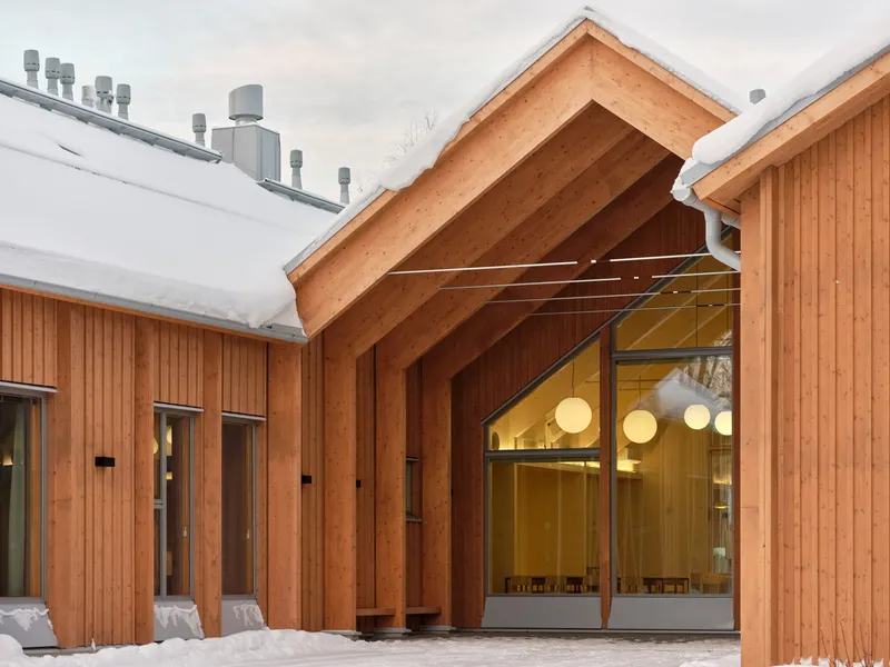 Timber gable entrance with exposed rafter structure and glazed doors under snow-covered roofs at dusk