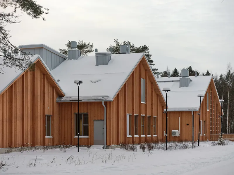 Street view of gabled timber volumes with vertical cladding and snow-covered roofs beside winter trees