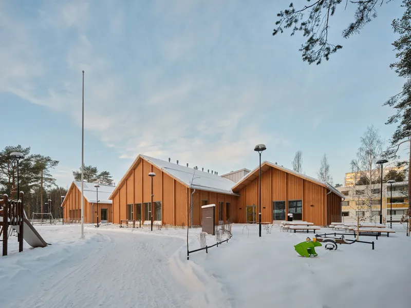 Cluster of timber-clad gabled volumes with playground equipment in a snow-covered clearing at twilight