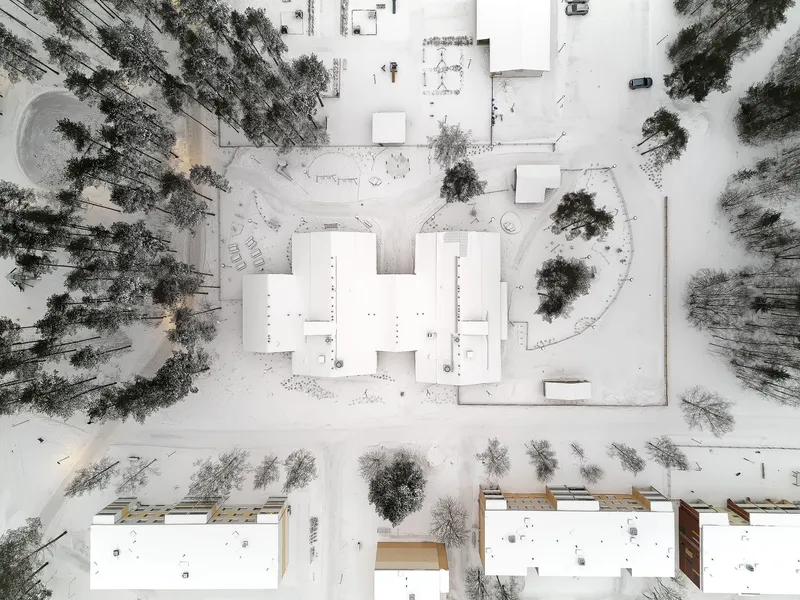 Overhead winter view showing white roofed volumes arranged around courtyards with scattered trees in deep snow