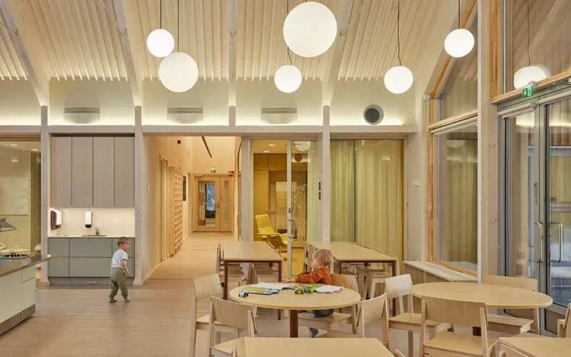 Interior playroom with blonde timber tables, globe pendant lights, and glazed walls opening to corridors beyond