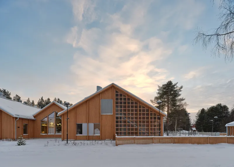 Timber gable with gridded translucent facade element dusted with snow at dusk beneath a streaked sky
