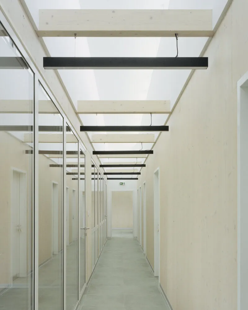 Long corridor with glass partitions and exposed timber beams punctuated by black pendant fixtures