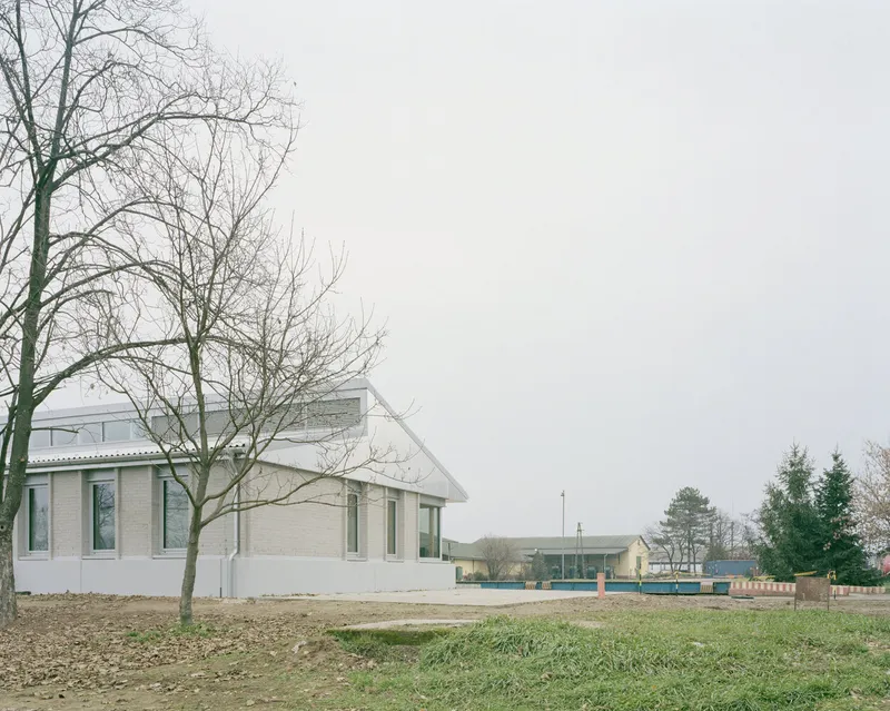 Gabled building with pale brick and corrugated roof beside leafless trees under grey sky