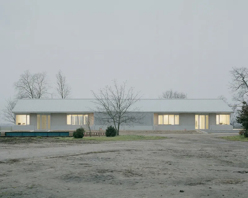 Long horizontal facade with punched window openings and corrugated metal roof across an asphalt parking area