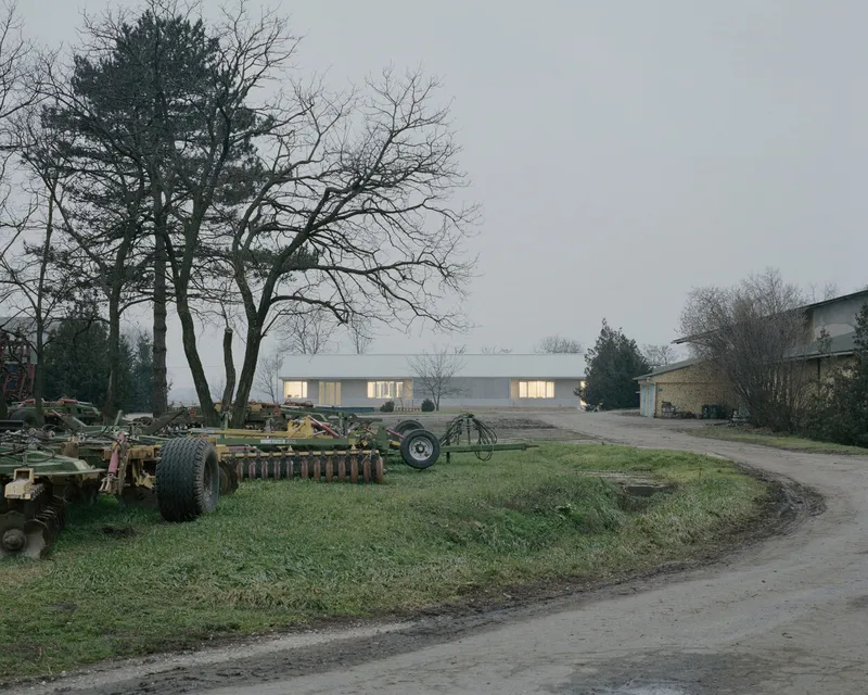 Linear low-slung building with illuminated windows and corrugated metal roof at dusk behind bare winter trees