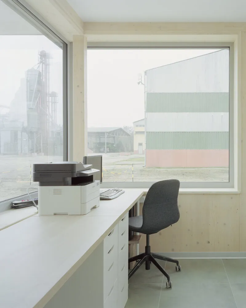 Office workspace with plywood walls and large corner windows overlooking industrial silos in fog