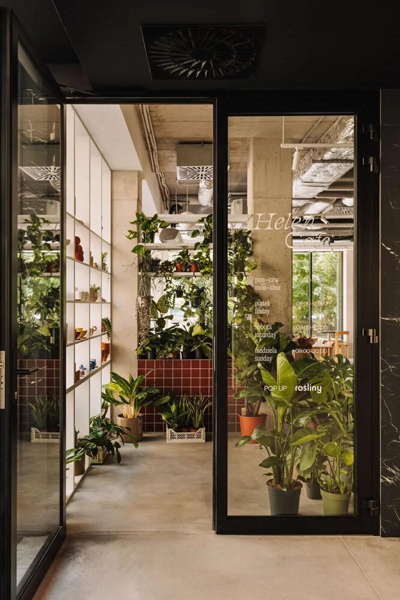 Glass storefront entrance with black steel frames revealing potted plants and white tile shelving within